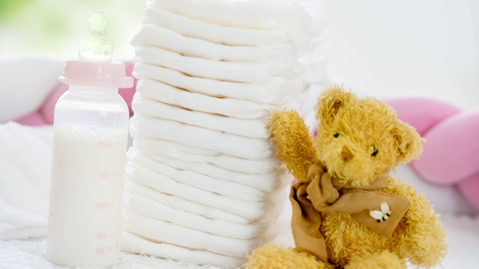 Soft yellow teddy bear next to stack of white diapers and baby bottle